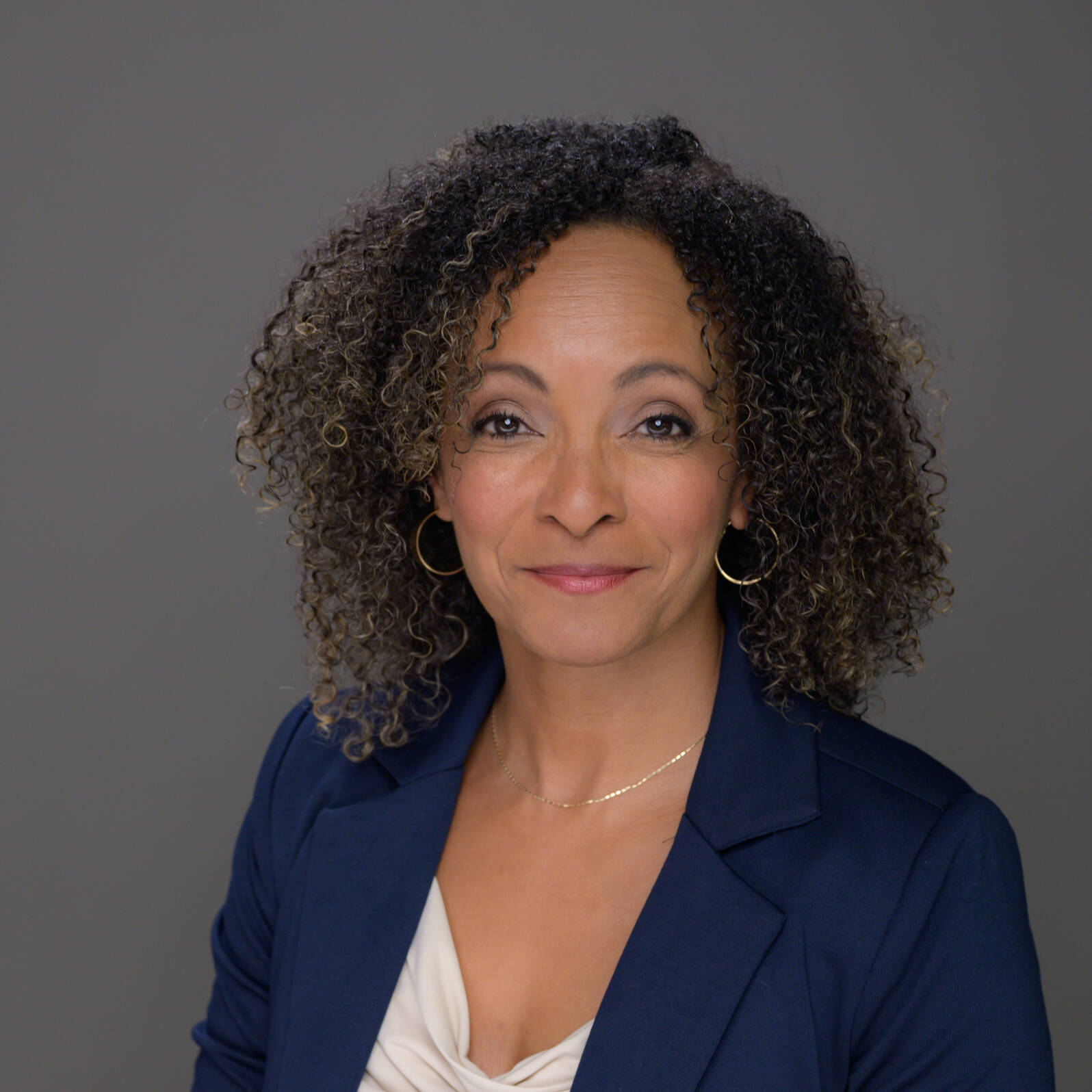 A person with curly hair wears a navy blazer over a white top and smiles gently against a neutral gray background.